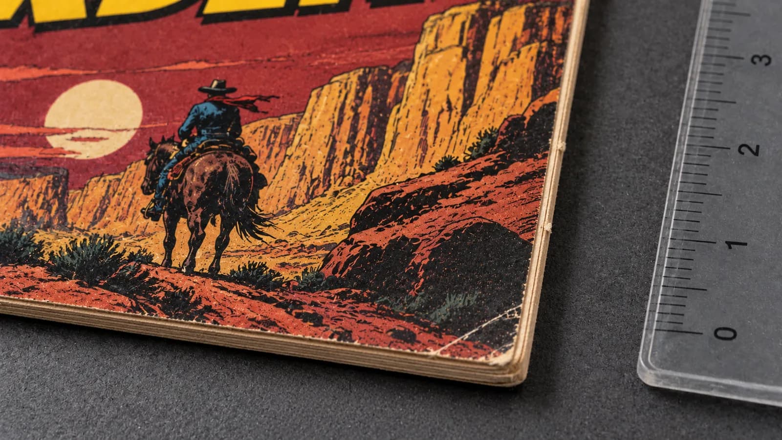 A western comic corner shows a small crease and pale edge wear beside a ruler on a dark inspection mat.